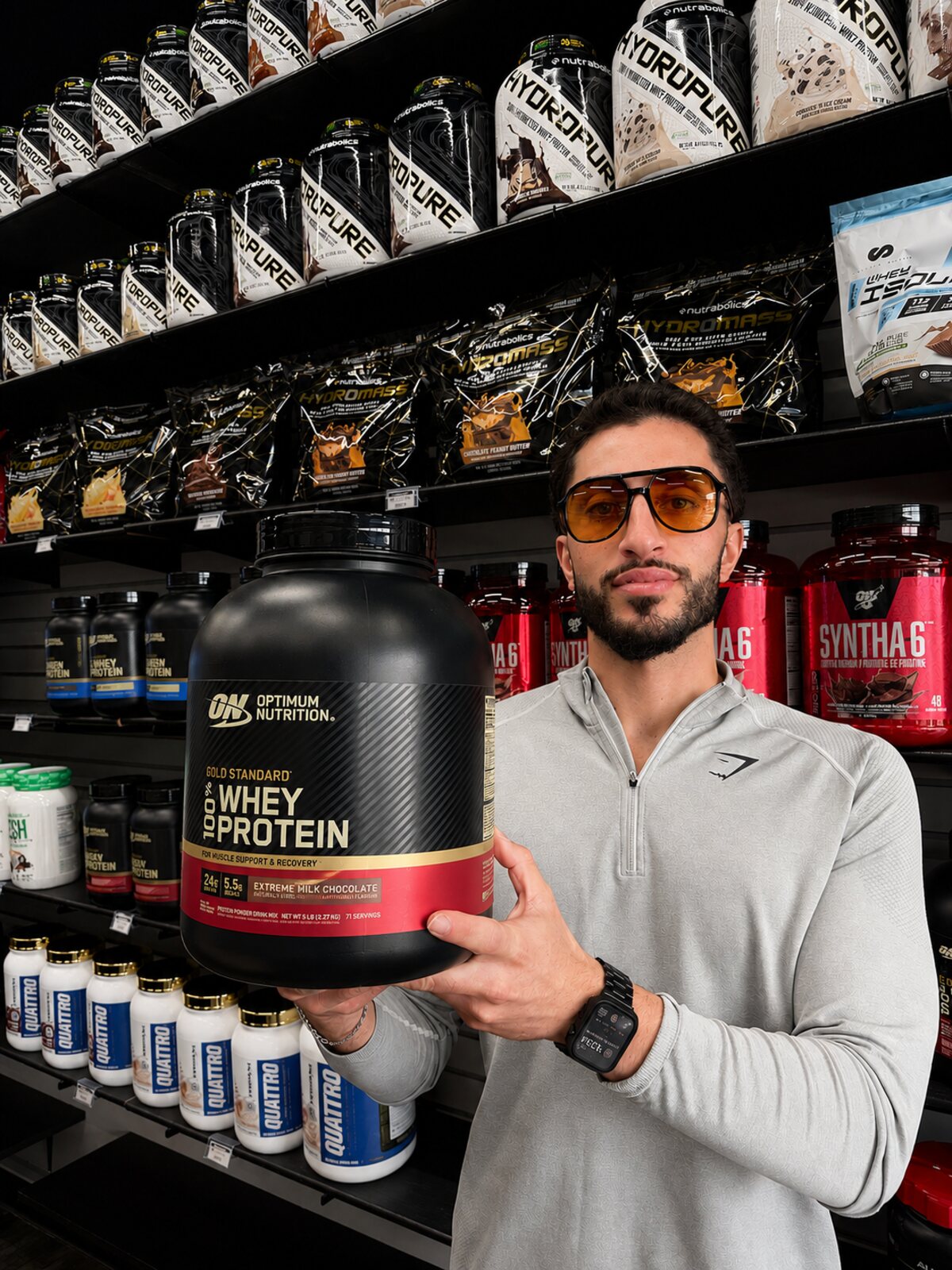 A Supplement World Newcastle team member holding a tub of Optimum Nutrition Gold Standard whey protein in front of the store's protein wall